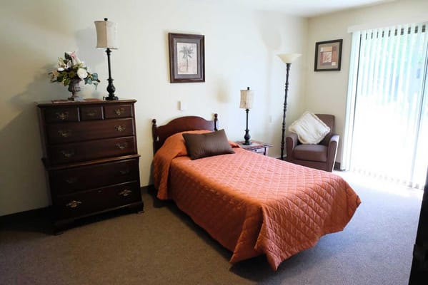 A cozy bedroom featuring a bed with orange bedding, a wooden dresser, lamp, and a chair.