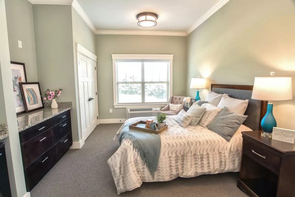 A well-decorated bedroom in a senior living facility with a bed, nightstands, and a window.