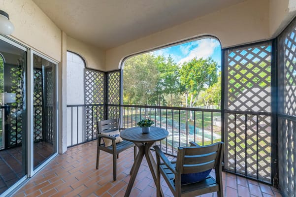 A small balcony with two chairs and a table, surrounded by lattice walls and greenery in the background.