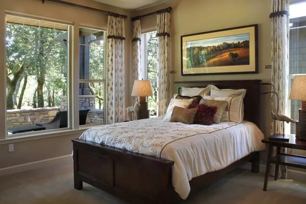 A well-decorated bedroom featuring a bed, lamps, and large windows overlooking trees.