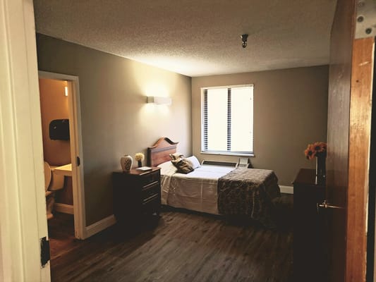 A well-lit bedroom in an assisted living facility with a bed, nightstand, and bathroom door visible.