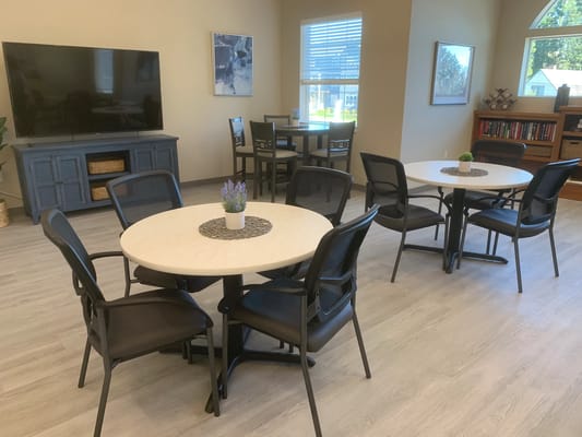 A bright common area with round tables and chairs, featuring a TV and bookshelves.