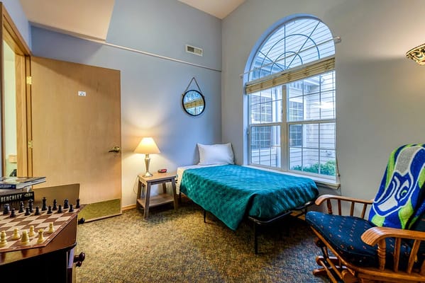 A well-lit memory care room featuring a bed, lamp, and armchair.