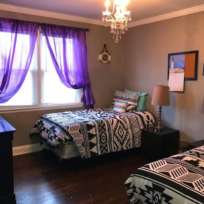 Two twin beds with patterned bedding and purple curtains in a bright room.