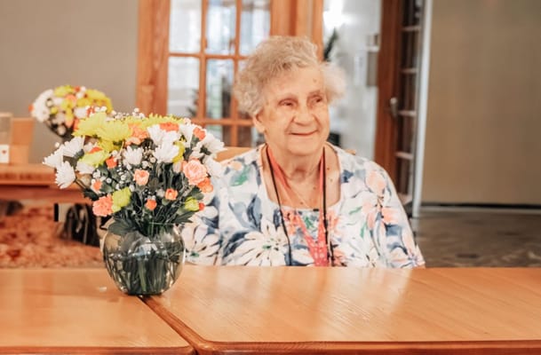An elderly woman sitting at a table with a floral arrangement