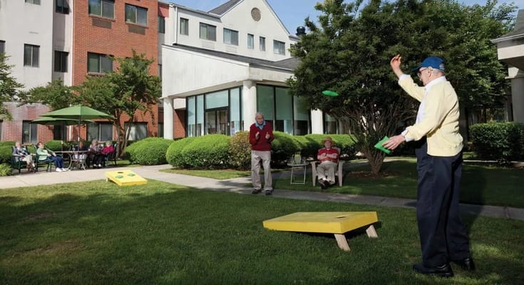 Senior residents playing cornhole outside in a green courtyard.