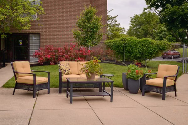 Outdoor seating area with tan chairs and a table surrounded by greenery and flowers.