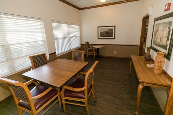 A cozy common area with a wooden table and chairs, featuring window blinds and decorative art.