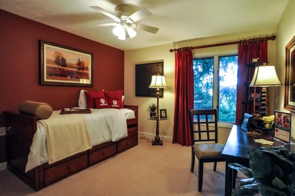 A cozy bedroom with a bed, desk, and large window in a senior living facility.