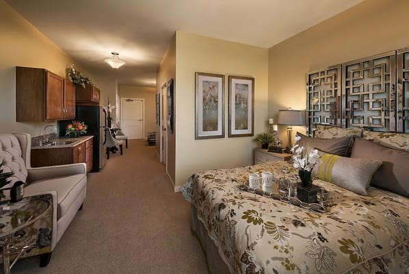 Private bedroom in an assisted living facility featuring a bed, kitchenette, and cozy decor.