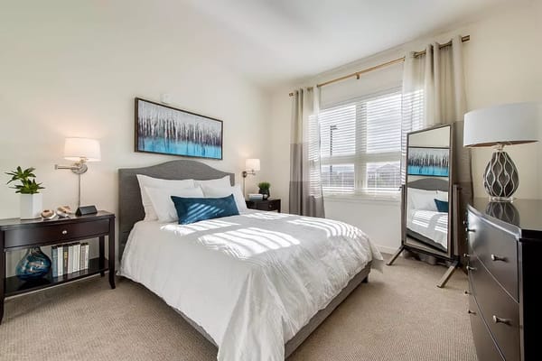 A bright bedroom featuring a bed, bedside table, and decorative mirror.