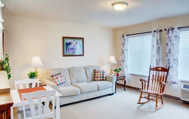 A comfortable living room with a beige couch, a wooden rocking chair, and large windows.