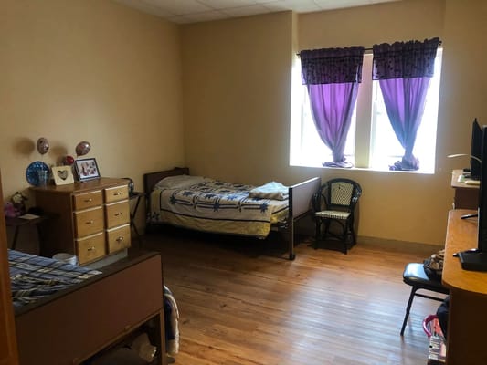 A bedroom with two beds and wooden flooring, featuring purple curtains and a dresser.