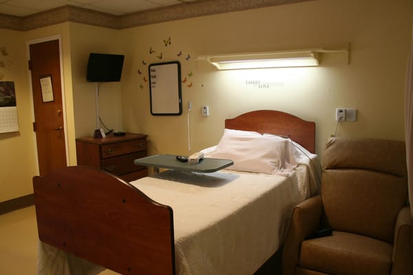 A cozy bedroom with a hospital bed, TV, and a chair at Steere House Nursing & Rehabilitation Center.