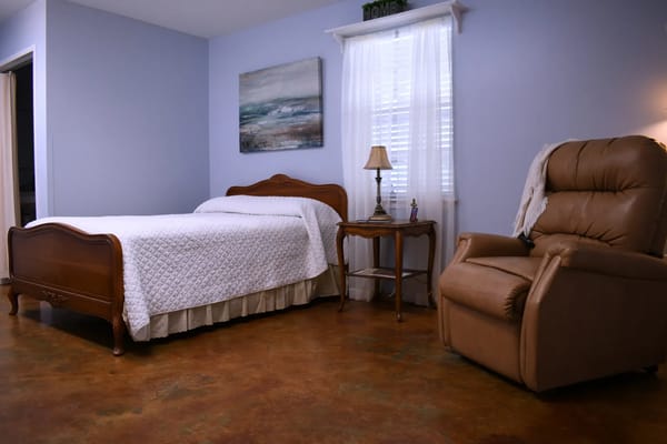 A cozy bedroom featuring a wooden bed, nightstand, lamp, and recliner.