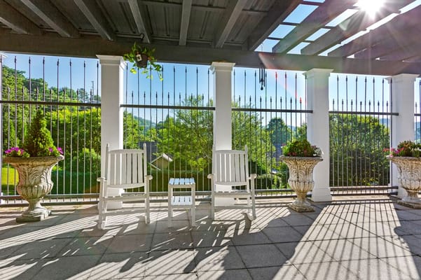 White rocking chairs and a small table on a sunny patio surrounded by greenery