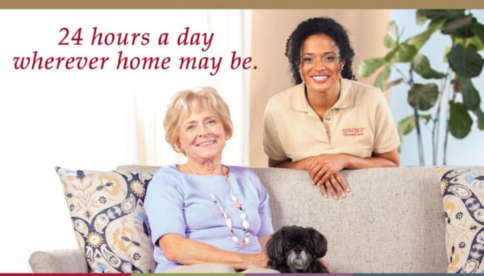 A caregiver and a senior woman smiling together on a couch with a small dog.