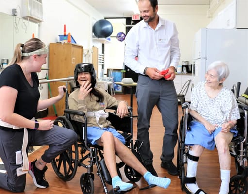 Two seniors in wheelchairs laughing with a staff member in a communal space.