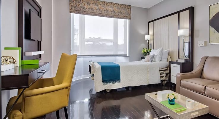 Cozy bedroom with a bed, armchair, and desk in a senior living facility.