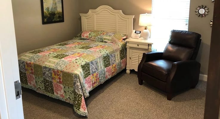 A well-decorated bedroom with a quilted bedspread, nightstand, and recliner.