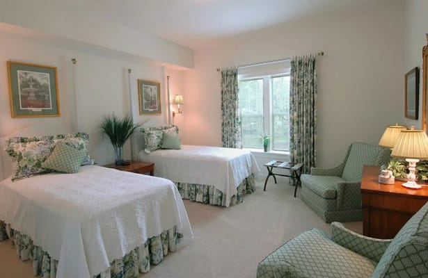 A cozy bedroom featuring two twin beds with floral bedspreads, a comfortable chair, and a window with light curtains.