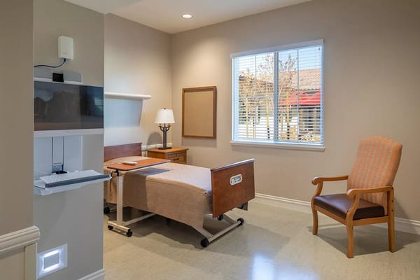 A well-lit patient room featuring a bed, chair, and window.