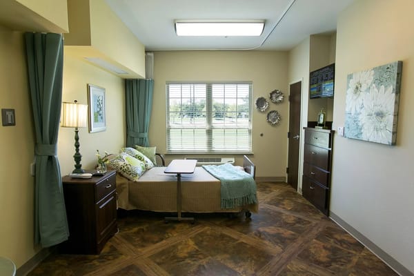 A well-decorated bedroom in a senior living facility with a bed, desk, and large window.