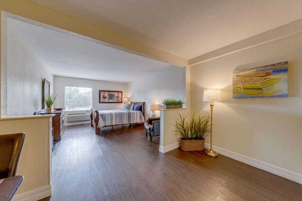 A well-lit, cozy bedroom with a bed and armchair, featuring a decorative painting and indoor plants.