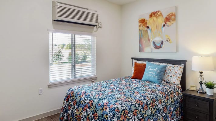 A cozy bedroom featuring a floral bedspread, lamp, and artwork