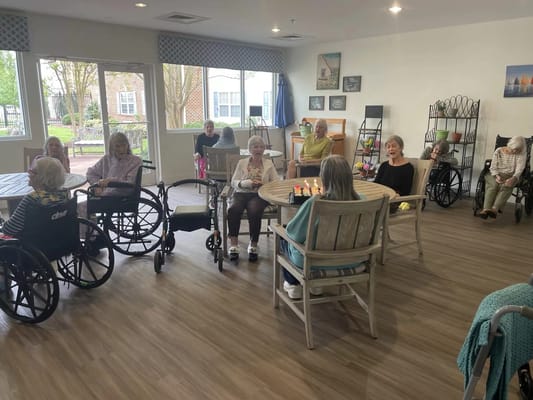 A group of elderly residents sitting in a common area, some in wheelchairs, engaging in conversation.