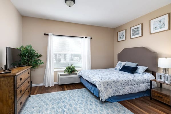 A neatly arranged bedroom featuring a bed, dresser, TV, and natural light.