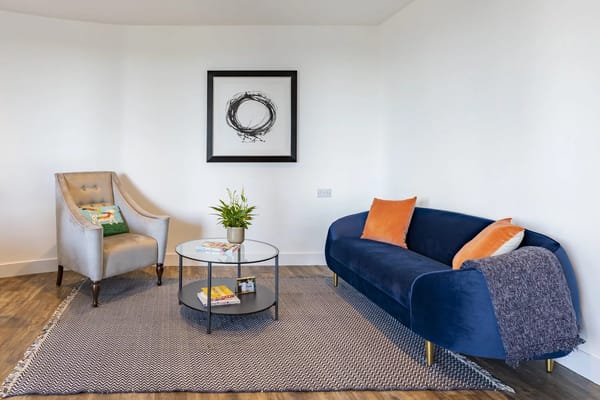 A stylish living room with a blue sofa, grey armchair, glass coffee table, and plant.