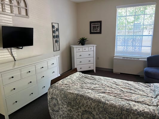 Brightly lit bedroom with a bed, dresser, TV, and seating area.