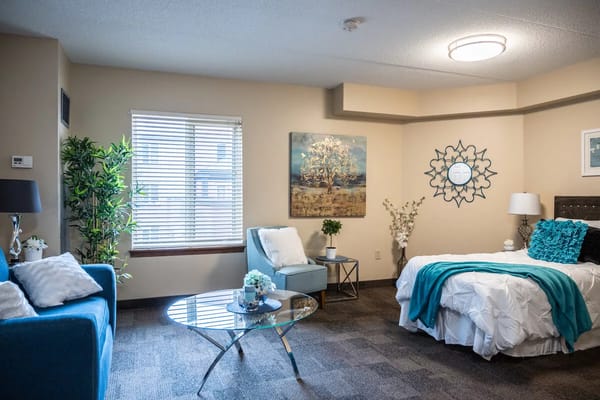 A well-lit bedroom featuring a bed with turquoise accents, a comfortable chair, and decorative plants.