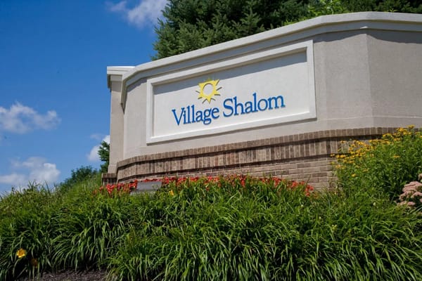 Sign for Village Shalom amid greenery and flowers