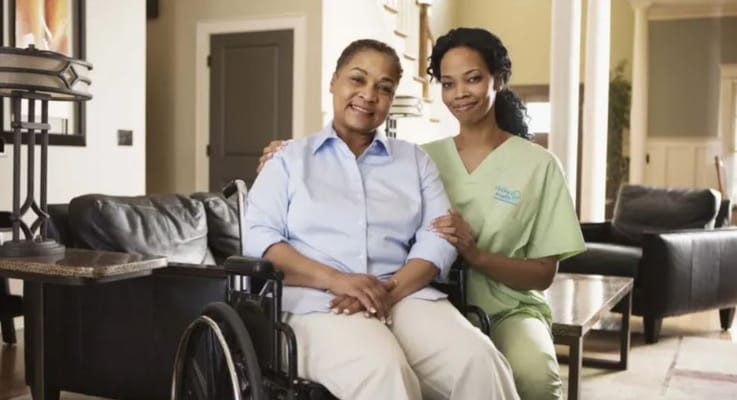 A caregiver with a smile sitting next to a senior woman in a wheelchair inside a cozy living space.