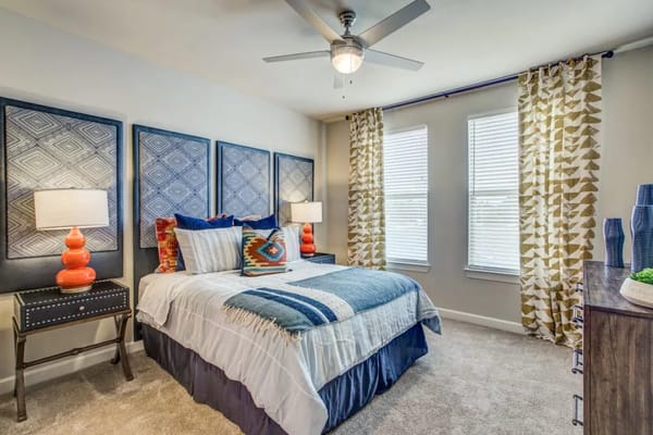 A styled bedroom with a bed, decorative pillows, lamps, and large windows.