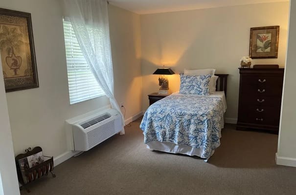 A well-furnished bedroom with a blue and white quilt, dresser, and a window with sheer curtains.