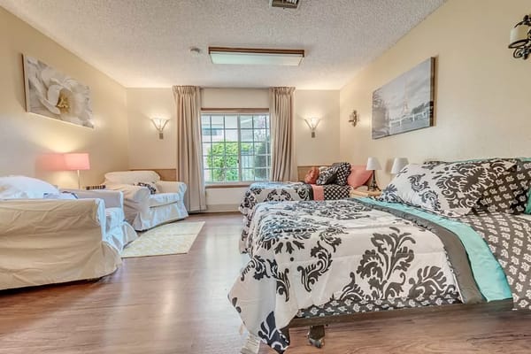 A well-lit resident room with two beds, armchairs, and a view outside.