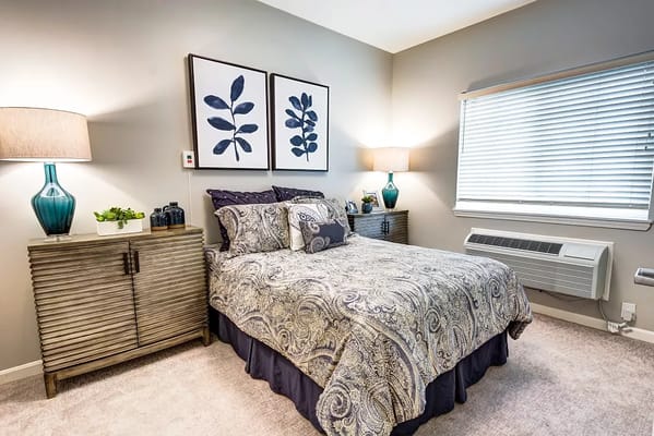 Cozy bedroom featuring a bed with patterned bedding, side tables with lamps, and a window with blinds.