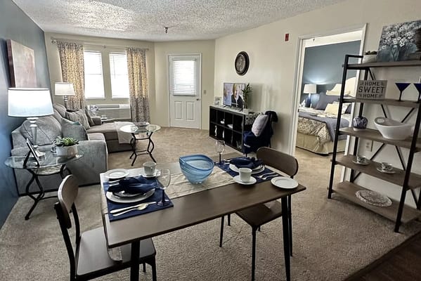 A bright, cozy living and dining area in a senior living facility with a dining table set.