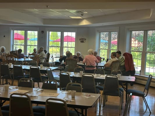 A group of seniors interacting in a dining area with large windows.