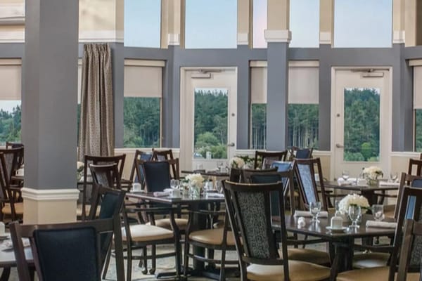 Spacious dining room with neatly set tables and large windows overlooking greenery.
