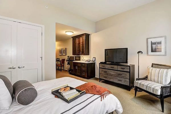 A spacious bedroom with a bed, TV, and armchair at Sunrise of Wilton.
