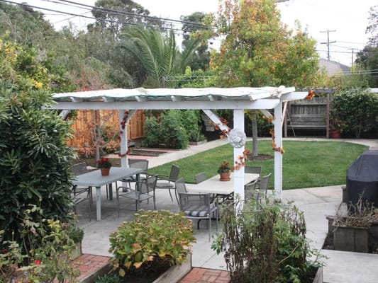 A serene outdoor patio with a pergola, tables, and chairs surrounded by greenery.
