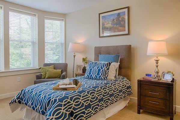 A well-decorated bedroom featuring a bed with a blue patterned blanket, a chair, and natural light from large windows.