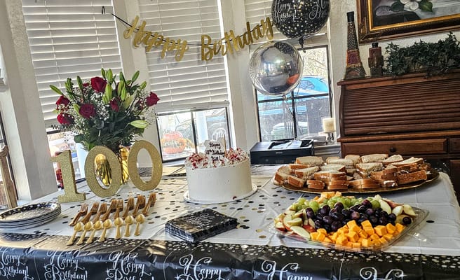 Table set for 100th birthday celebration with cake, food, and flowers