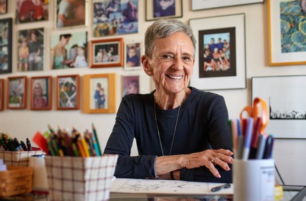 Senior woman smiling while working on art with colorful supplies around her.
