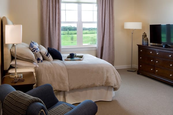 A well-furnished bedroom with a bed, lamp, TV, and window view.