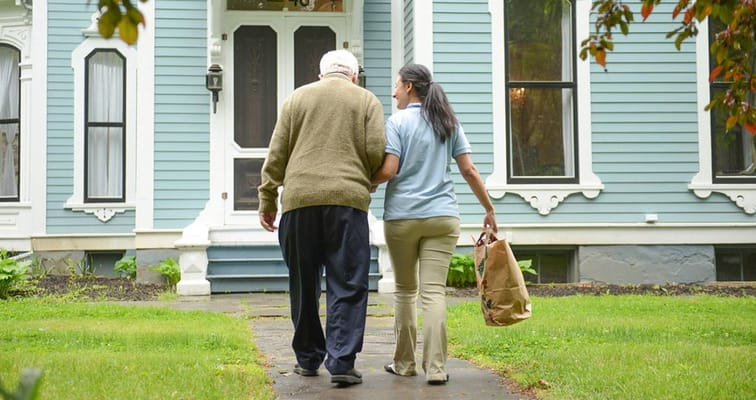 A caregiver accompanies a senior man walking towards a house.